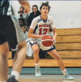 Basketball player in white jersey dribbles on court during a game, with spectators in bleachers behind.