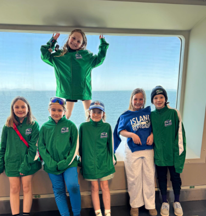 Five children in green jackets pose indoors by a large window overlooking the ocean.