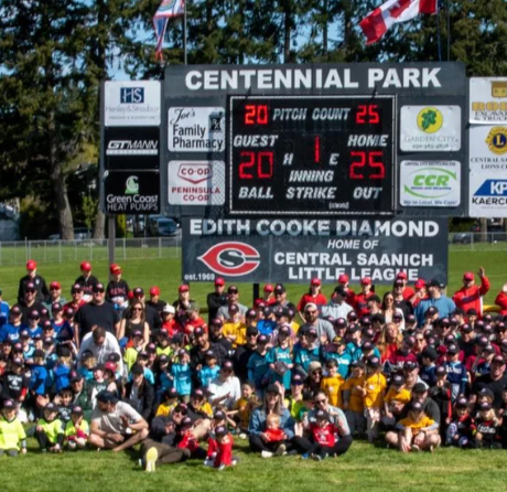 Baseball team group photo at Centennial Park scoreboard, with many players in red, yellow, and blue uniforms