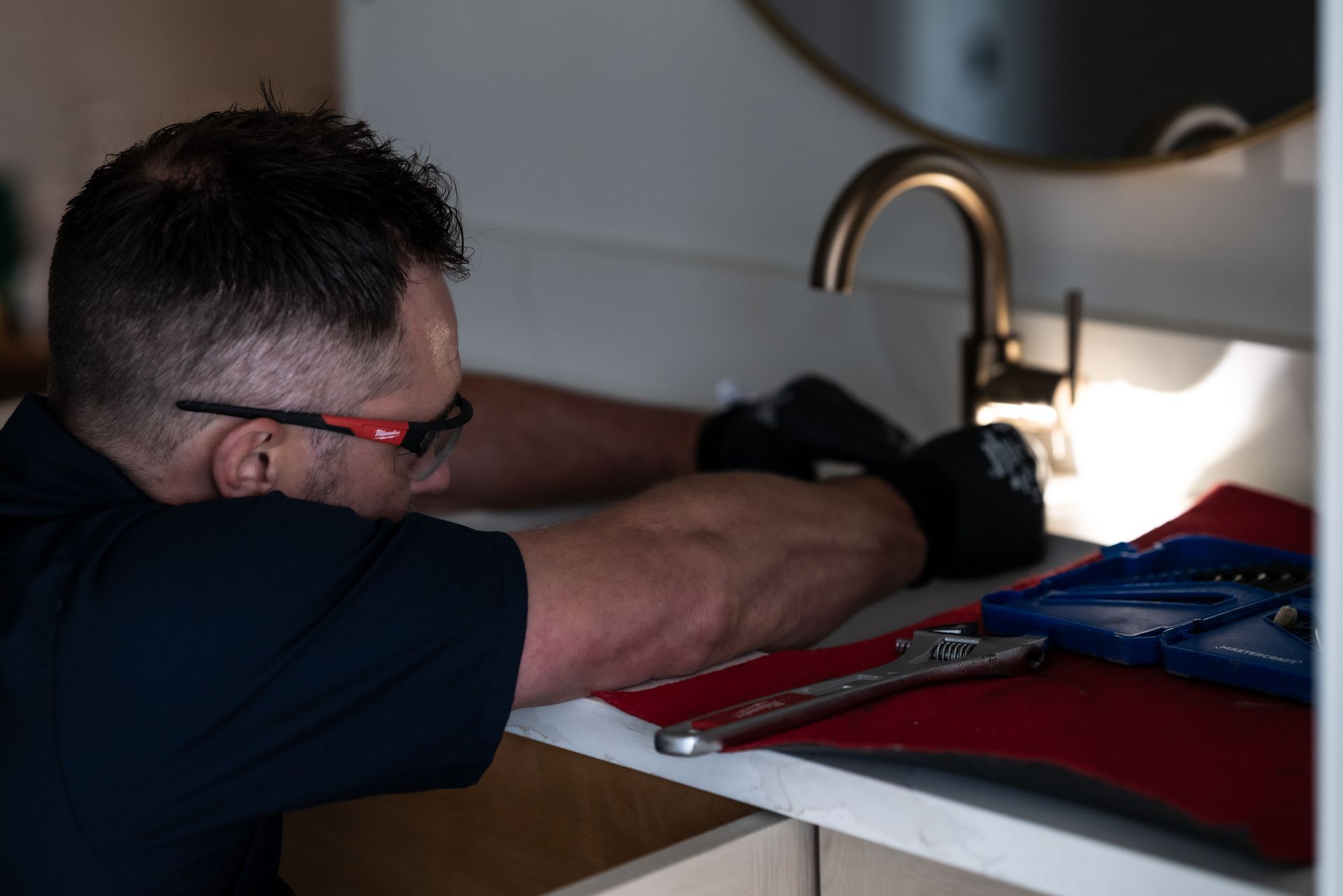 Plumber tightening a pipe under a sink with a wrench, wearing gloves.