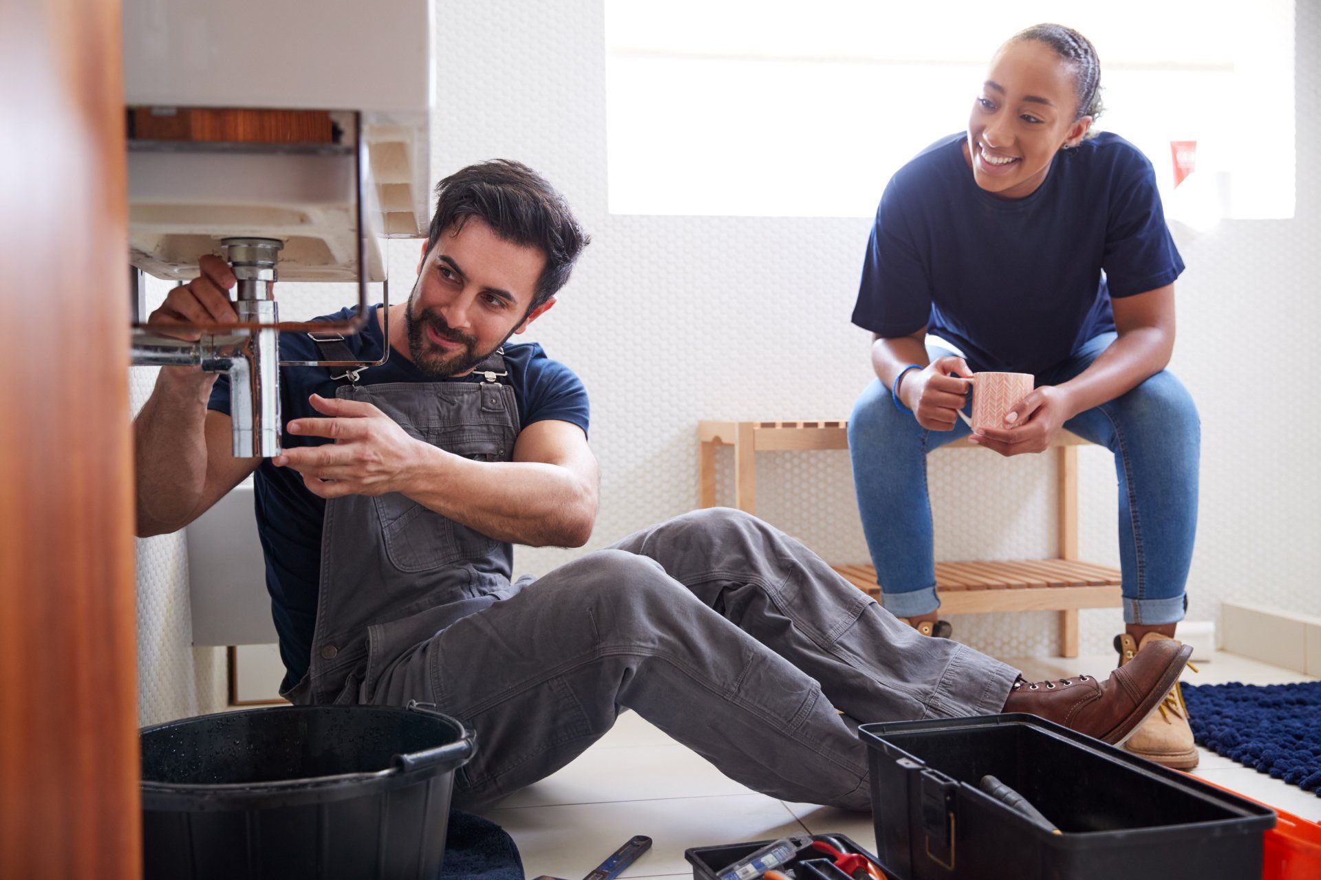 A man is sitting on the floor fixing a sink while a woman sits on a stool.