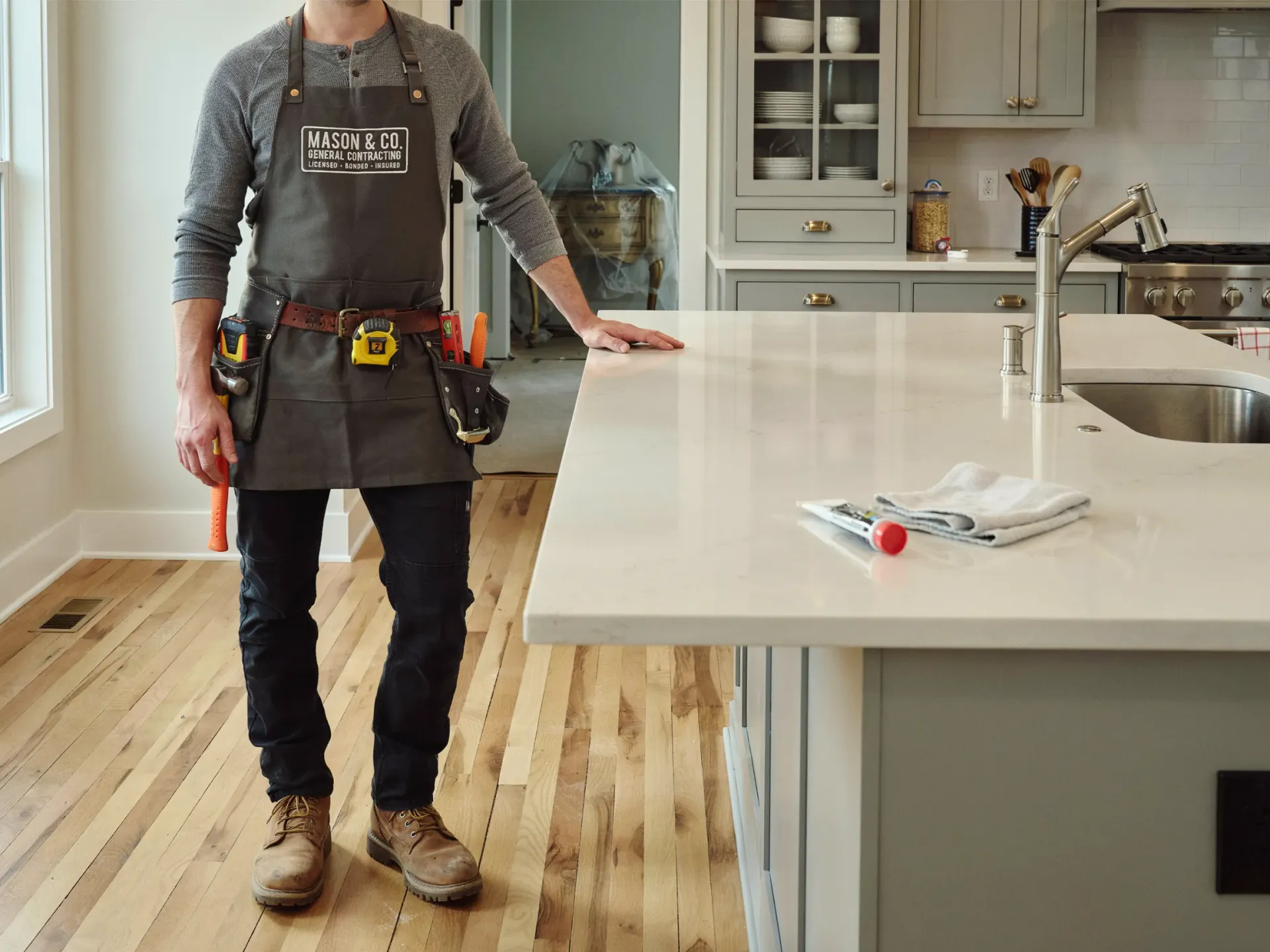Person in a kitchen wearing a tool belt beside a white island with a screwdriver on top
