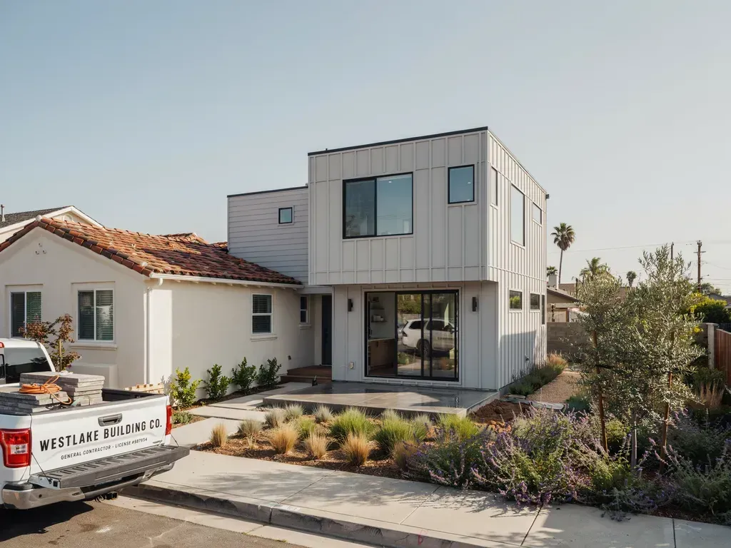 Modern two-story white house under construction beside a older single-story home, with landscaping in front.