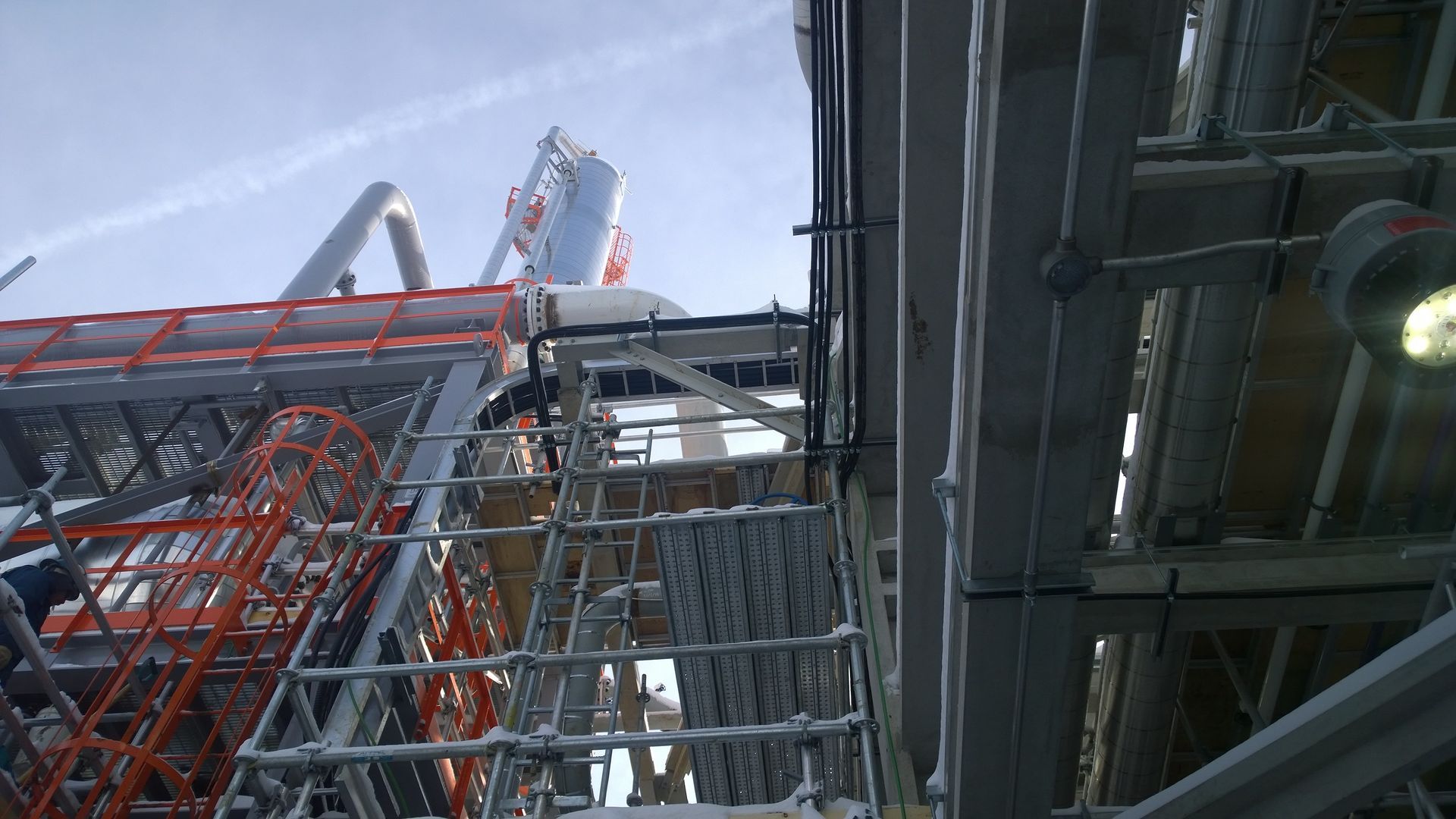 Industrial facility with red and gray metal structures, viewed upward against a cloudy sky.