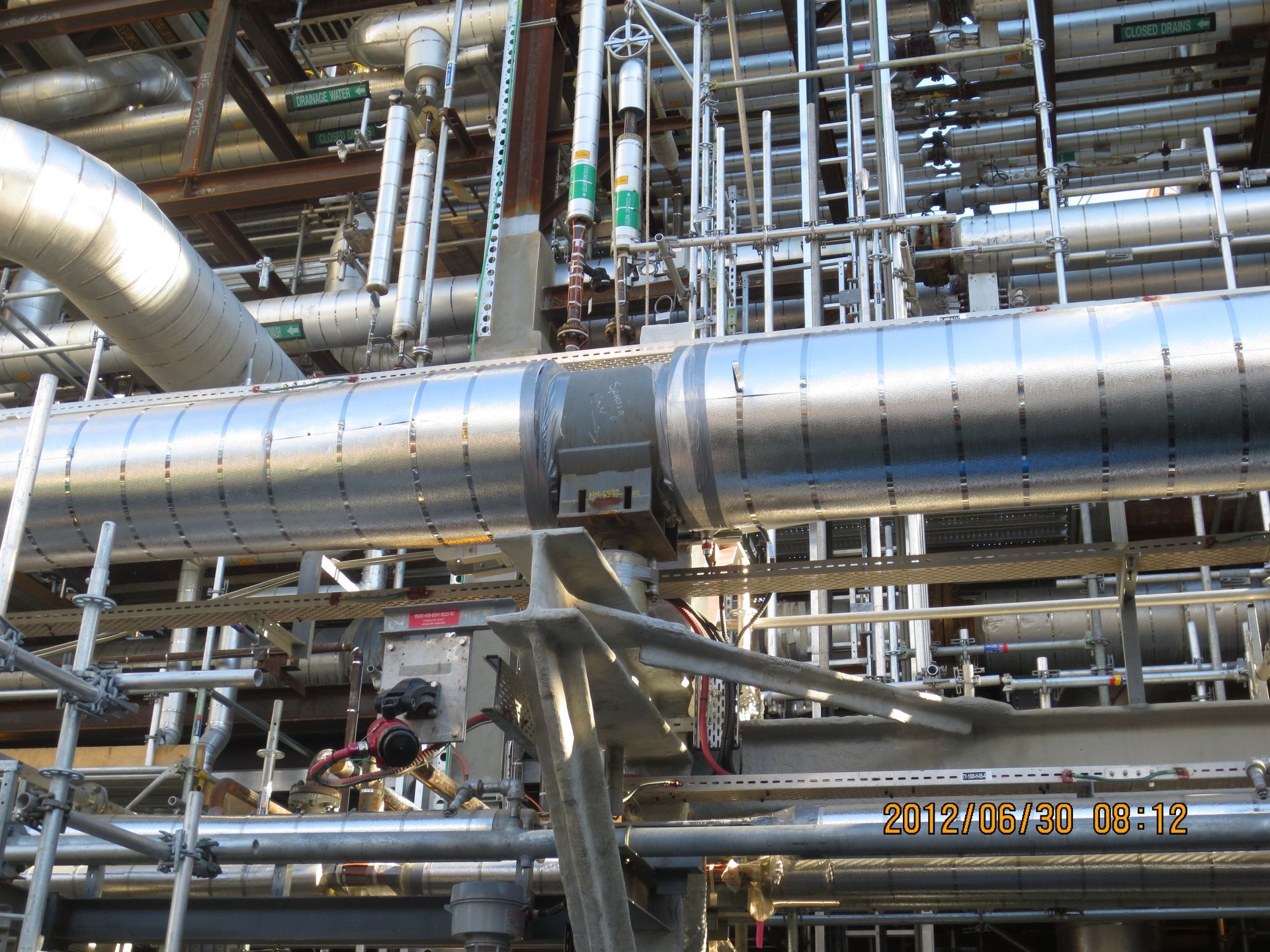 Industrial pipes and metal catwalks in a dense plant setting, with workers below.