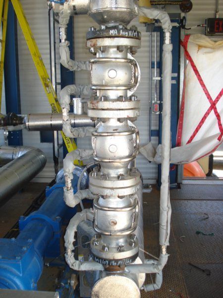 Vertical industrial pipe assembly with stacked metal valves and blue machinery in a factory setting