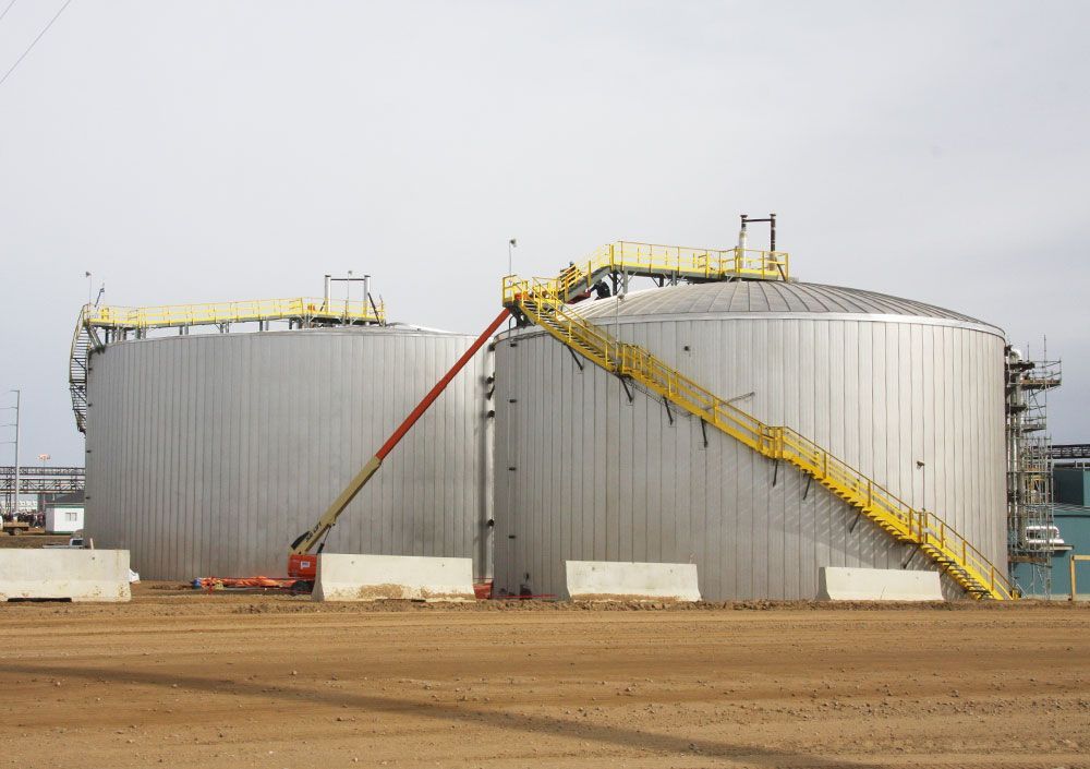 Two large industrial grain silos with yellow catwalks and a red conveyor in a dirt lot under a gray sky