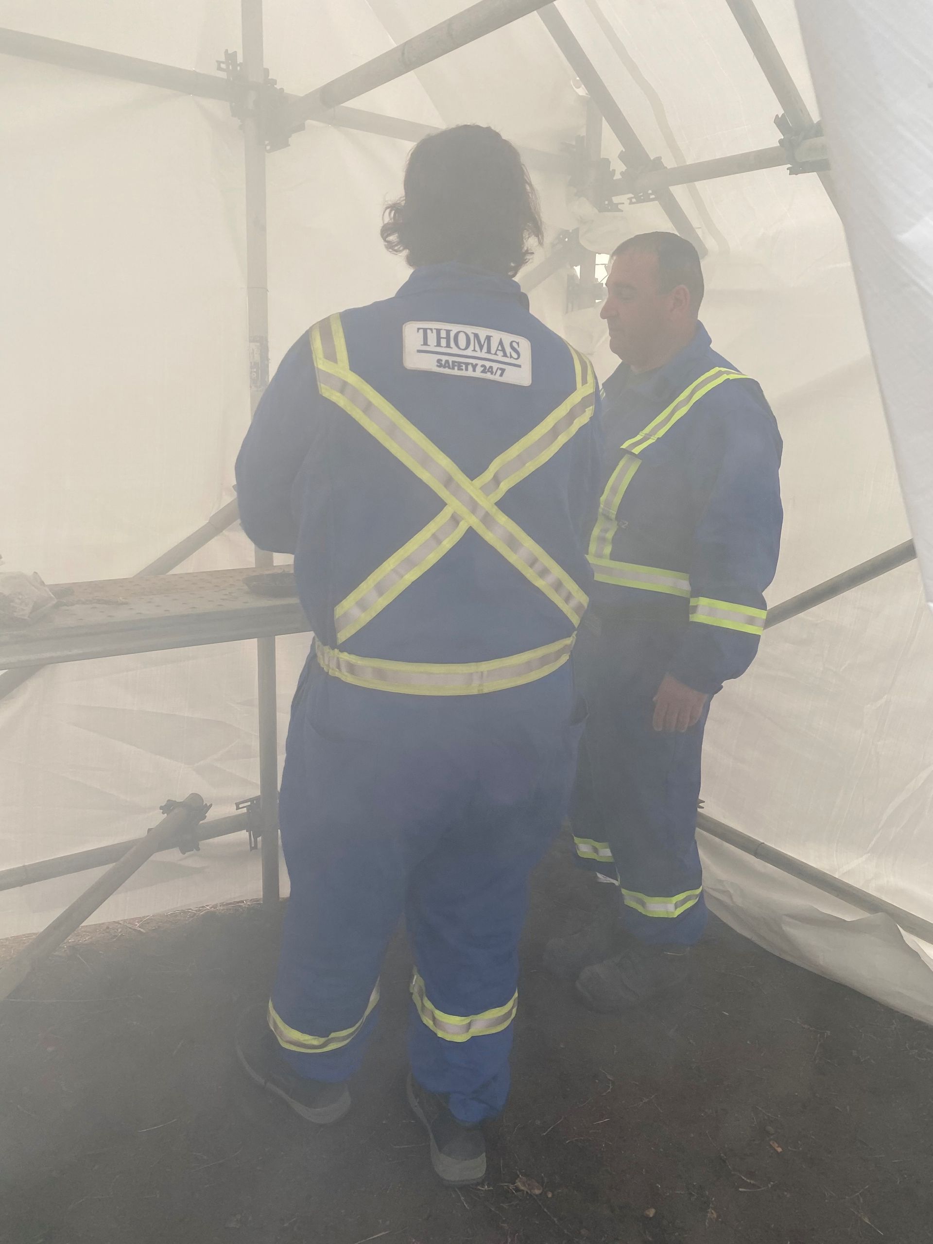 Two workers in blue safety gear with reflective stripes inside a white tent-like structure.