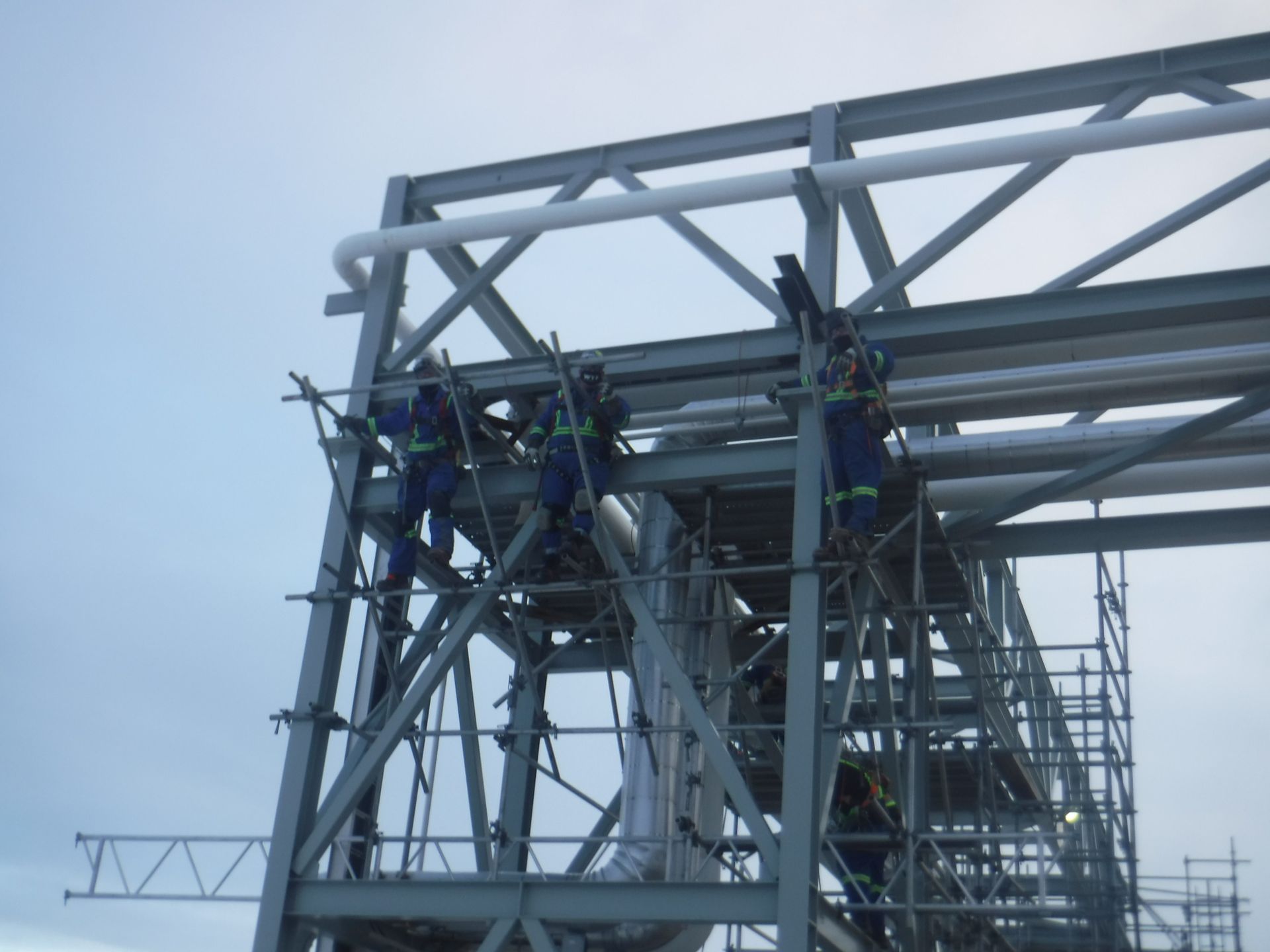 Workers on a metal structure, using harnesses and scaffolding against a cloudy sky.