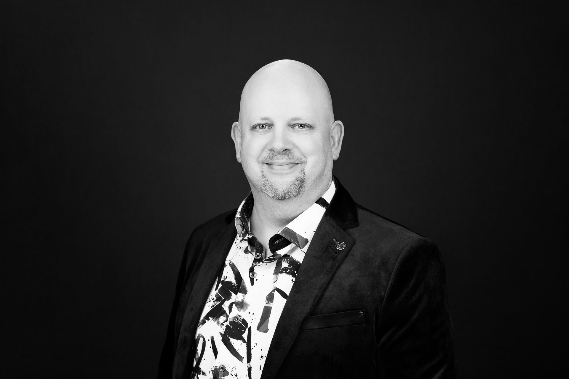 Smiling bald man in a dark blazer and patterned shirt against a black background
