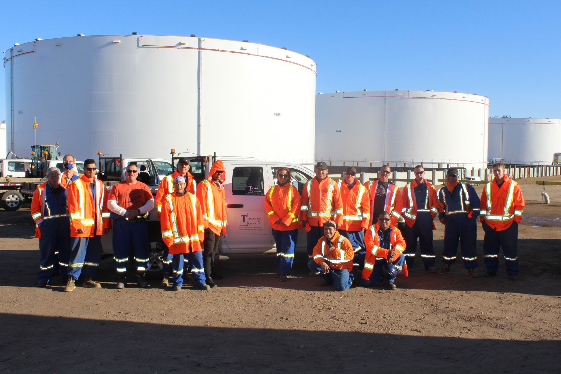 Workers in orange safety vests posing in front of large white storage tanks on an industrial site