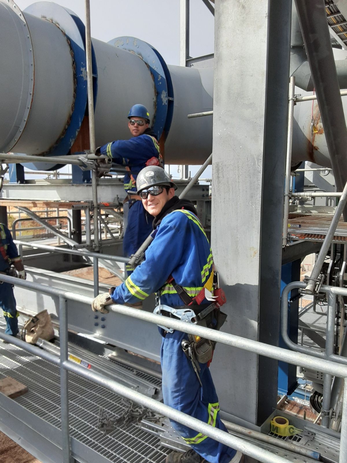 Two workers in blue safety gear stand on a metal platform beside large industrial pipes.