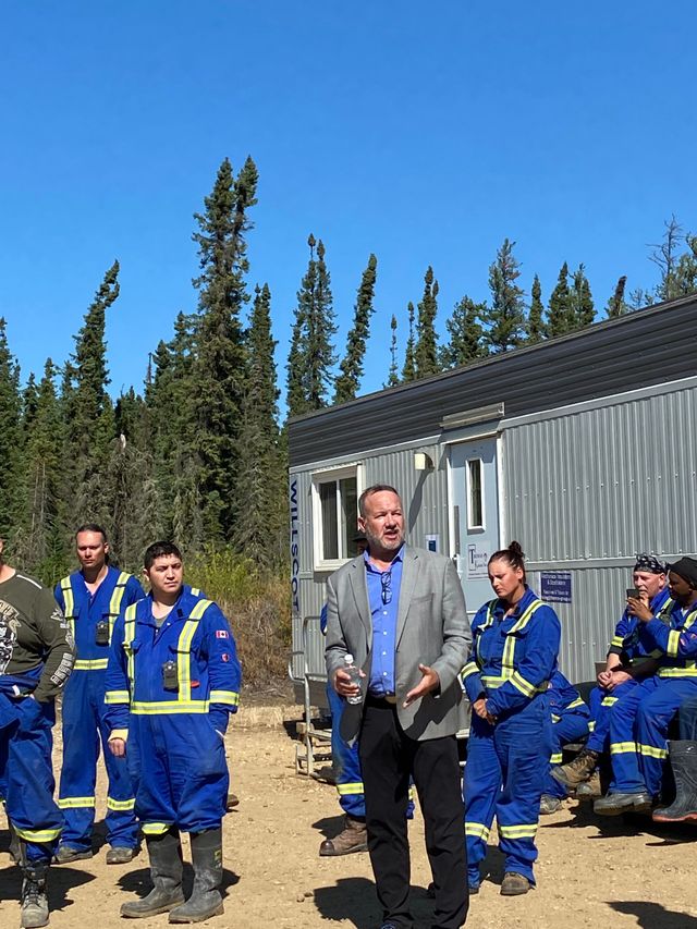Workers in blue coveralls gathered beside a metal building outdoors, with one man standing in front.