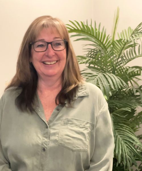 Smiling person in glasses and a light green shirt standing indoors beside a leafy plant.