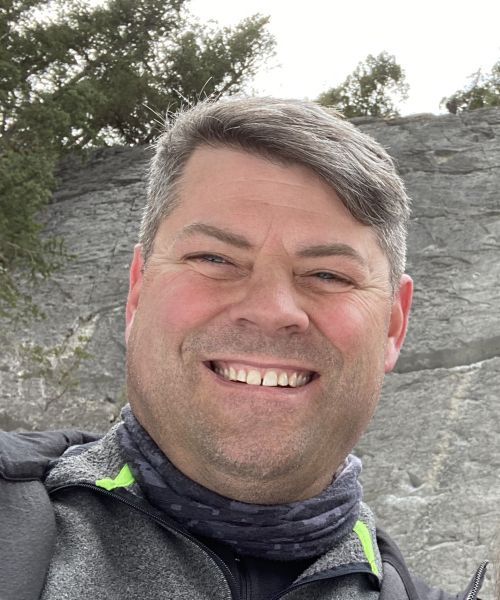 Smiling man in a dark jacket and neck gaiter outdoors in front of a rocky cliff.