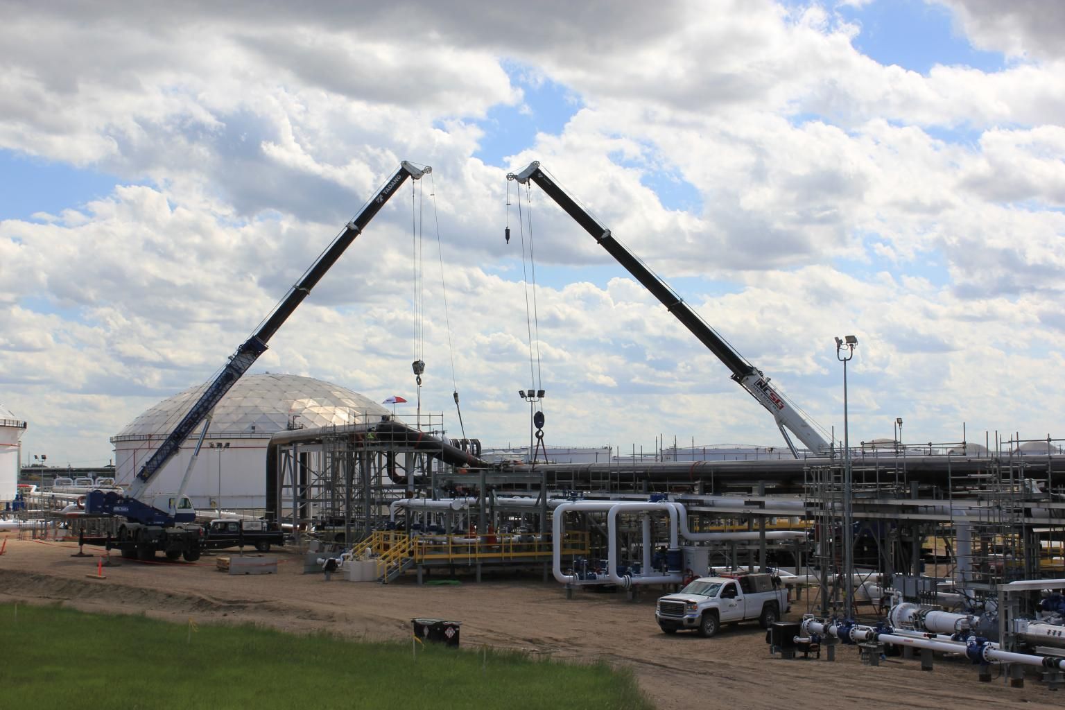 Two cranes lifting steel beams at an industrial construction site under a cloudy sky.