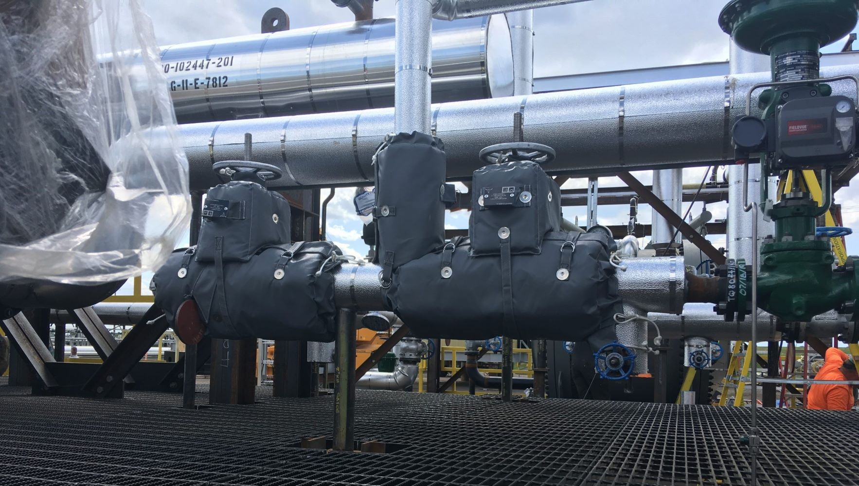 Industrial piping and valves on a platform, with workers in orange safety gear in the background