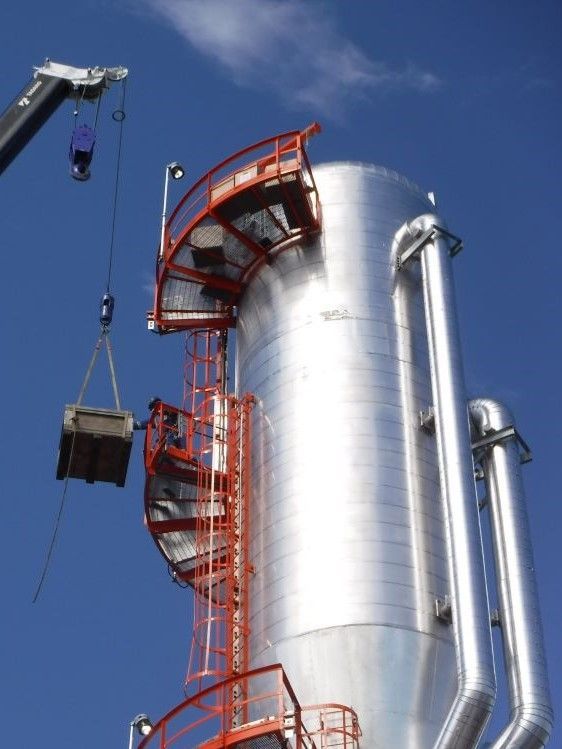 Crane lifting a large metal tank beside a shiny industrial silo with red stairways under a blue sky
