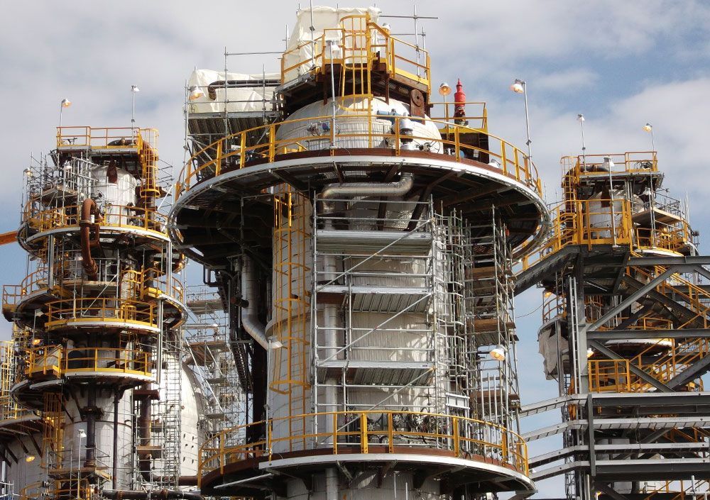 Large industrial structure under construction, wrapped in scaffolding and yellow safety railings.