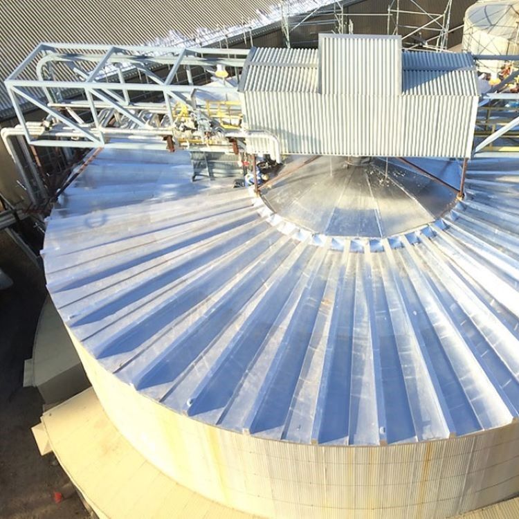 Aerial view of a large silver corrugated industrial silo with a metal conveyor structure attached.