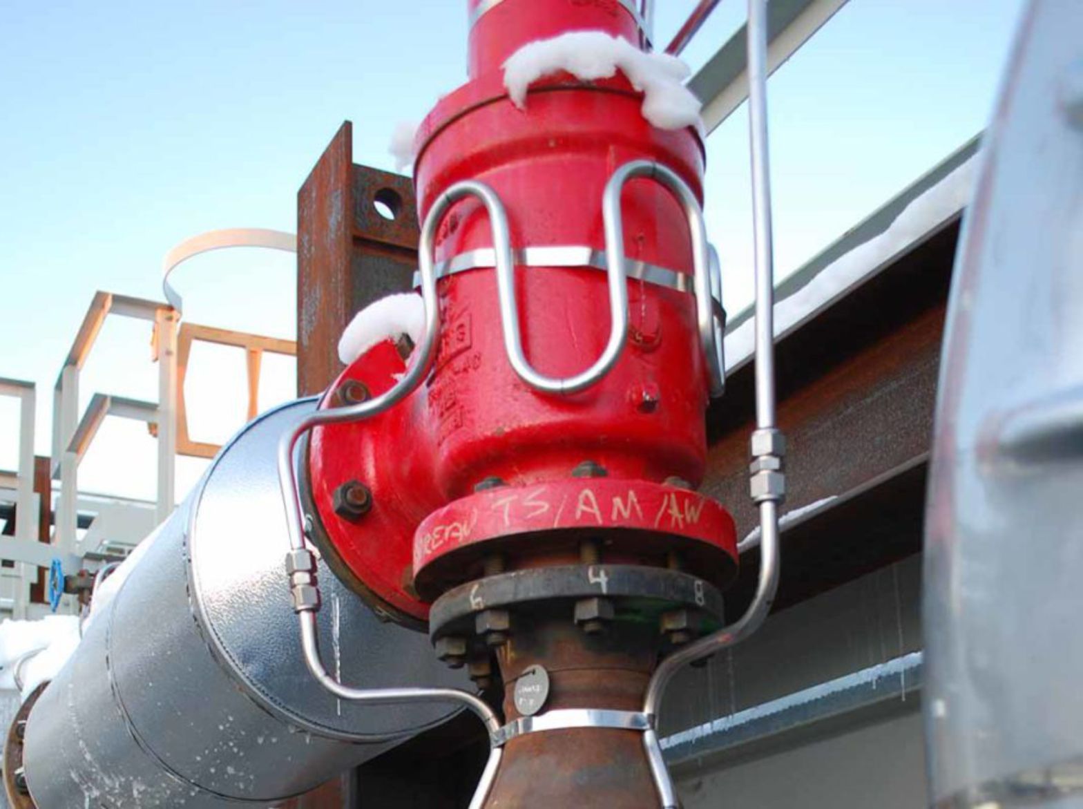 Red industrial valve with snow on top, mounted on metal piping outdoors.