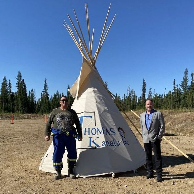 Two people standing beside a tan tipi in a sunny open field with blue sky and trees.