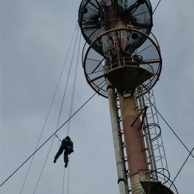 Person rappels beside a tall industrial tower with large circular fans against a cloudy sky