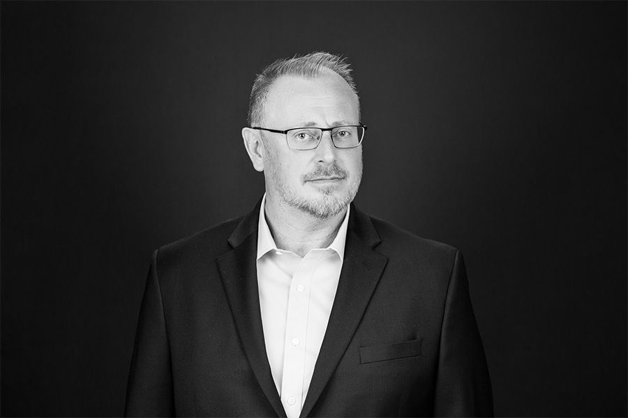 Man in a dark suit and glasses standing against a dark background, looking slightly to the side.