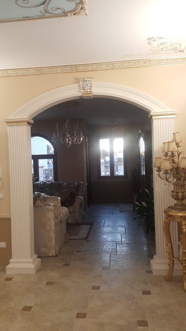Porta ad arco che conduce a un soggiorno poco illuminato con pareti color crema, pavimento piastrellato e lampadario visibile dall'interno.