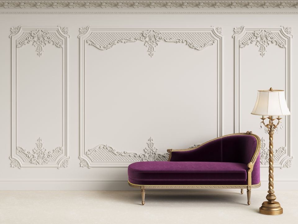 Elegante parete bianca con modanature decorate, una chaise longue viola e una lampada da terra dorata.
