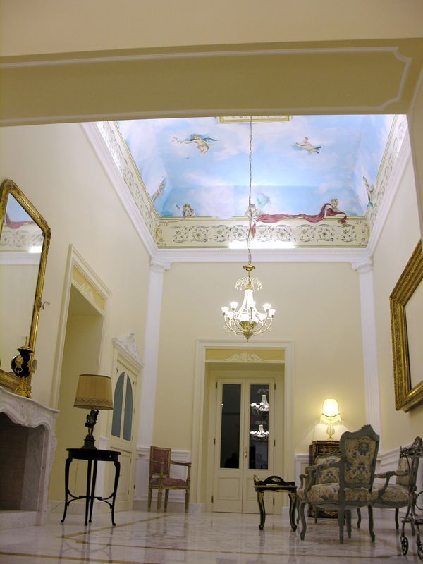 Elegante stanza color crema con lampadario, soffitto dipinto color cielo, camino e sedie antiche.