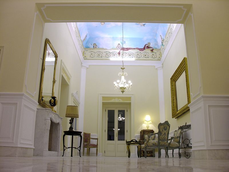 Elegante corridoio bianco con lampadario, sedute decorate, specchi e un affresco sul soffitto.
