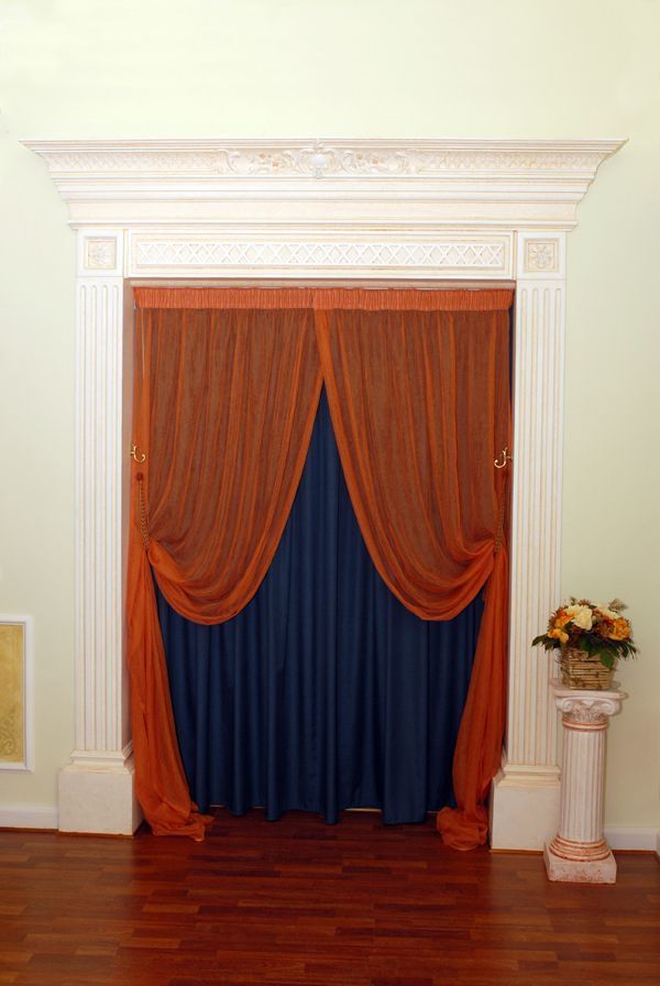 Portale decorato con drappeggi arancioni e blu, fiancheggiato da colonne bianche e una composizione floreale.