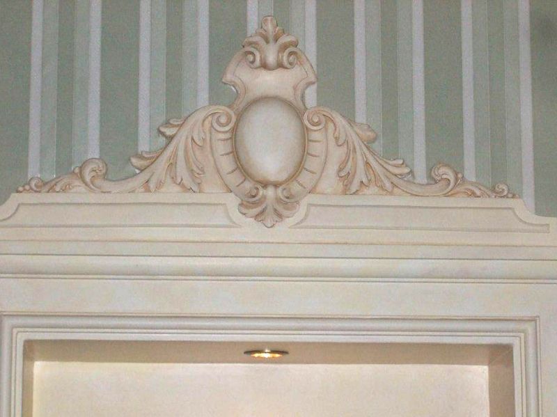 Sopra una porta, su uno sfondo di carta da parati a righe verdi, si erge una cornice decorativa color crema con intagli a volute.