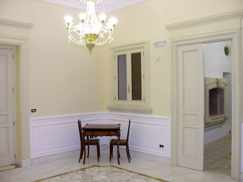 Stanza vuota color crema con lampadario, tavolino e sedie, boiserie bianca e porta aperta verso un'altra stanza.