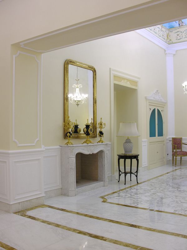 Elegante corridoio color crema con pannelli bianchi, finiture dorate, camino, specchio e lampadario.