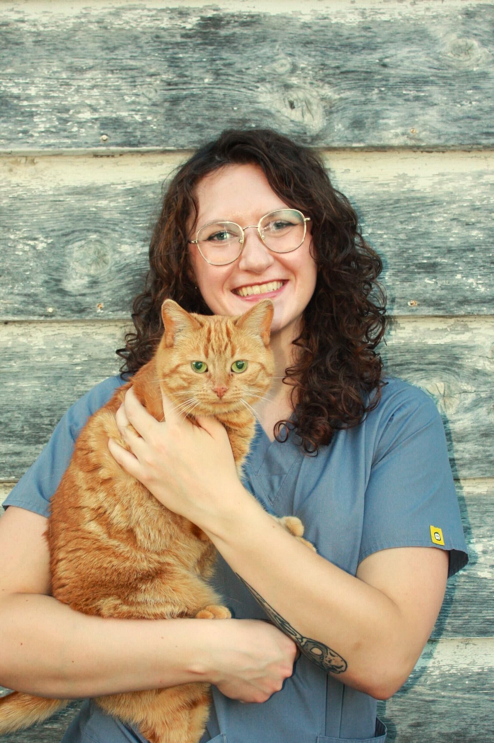 Woman in scrubs holds an orange cat against a weathered wooden wall, both smiling.