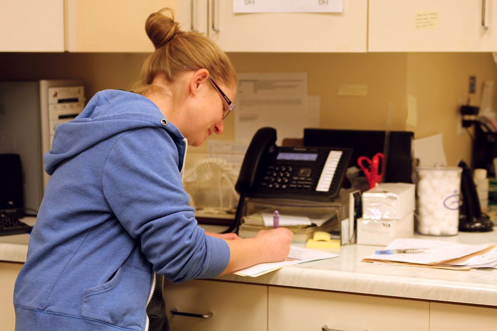 Woman in blue hoodie writing at desk with phone and paperwork.