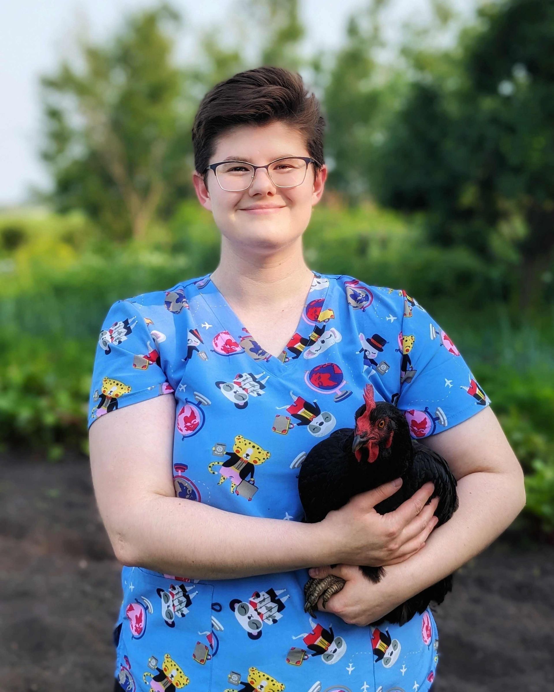Woman in blue scrubs holds a black chicken outdoors; smiles at the camera.