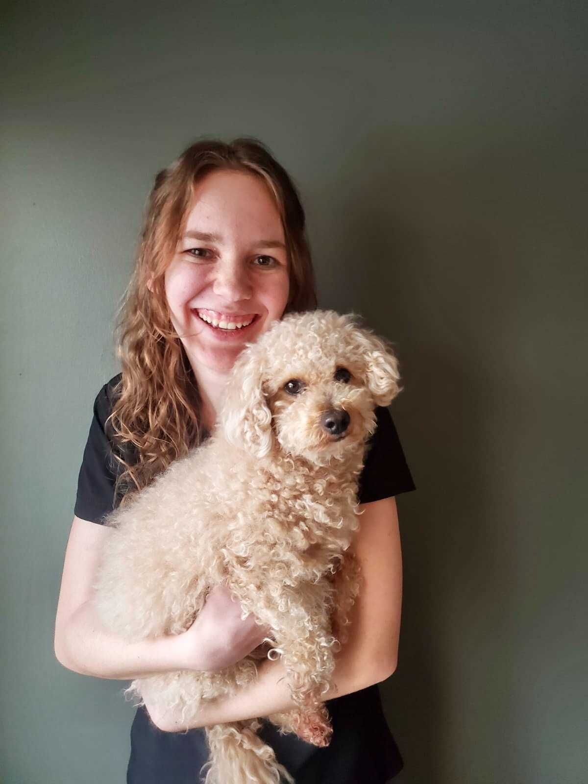 Woman smiling, holding a light brown poodle, standing in front of a green wall.