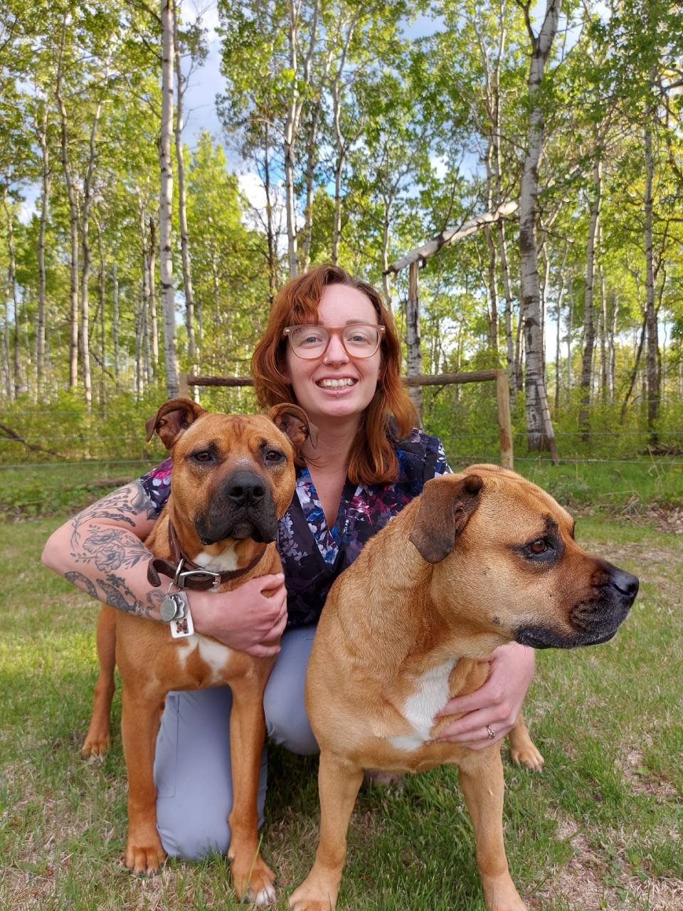 Woman kneels, smiles, and hugs two tan dogs in a grassy area with trees in the background.