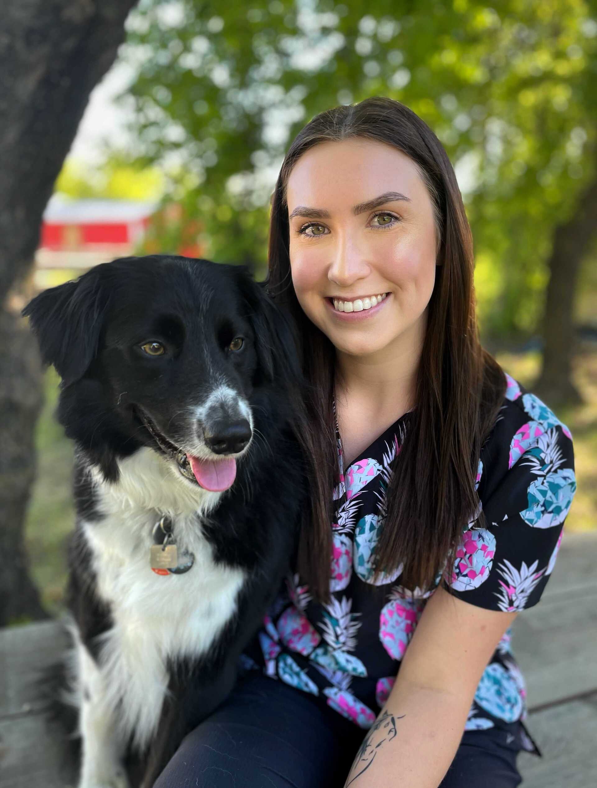 Woman in pineapple print scrubs smiles next to a black and white dog outdoors.