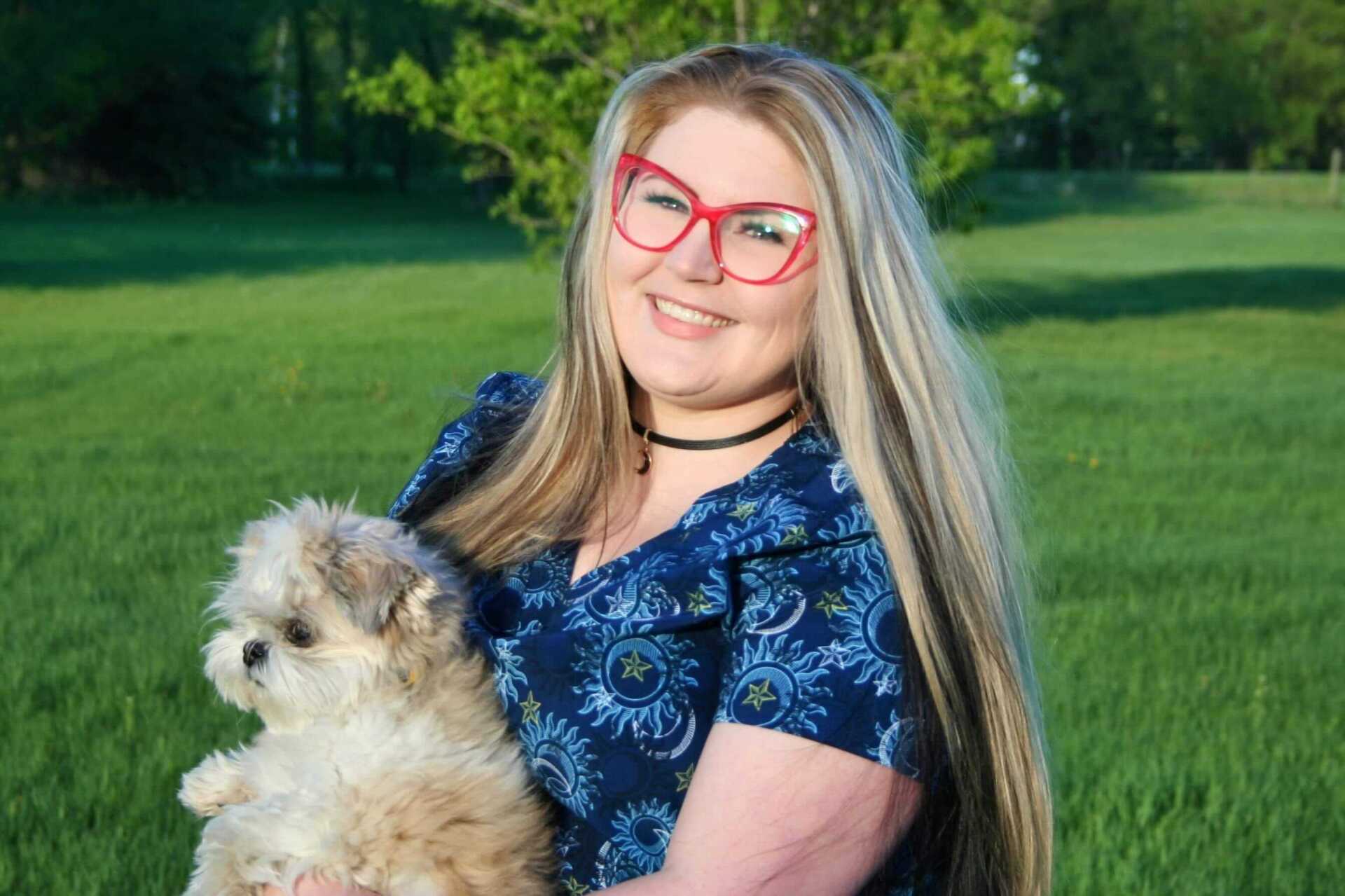 Woman with long blonde hair, red glasses, and blue floral shirt, holding small fluffy dog in grassy field.
