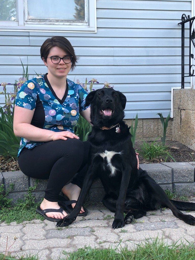 Woman in patterned scrubs sits with black dog on brick patio.