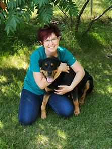 Woman in blue shirt embraces a black and brown dog in a grassy, outdoor setting.