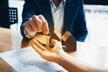 Person handing keys to another person, near a document and a small wooden house.