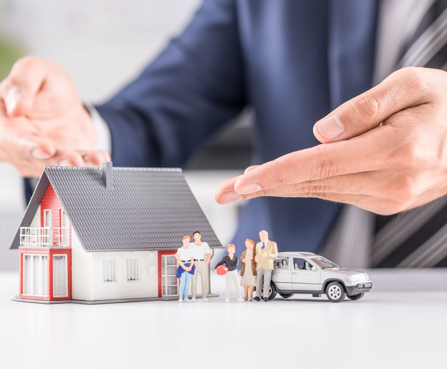Hands of a person protecting a model house, family figurines, and a car; likely insurance-related.