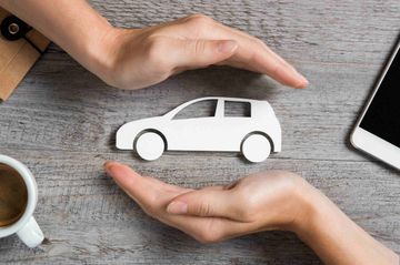 Hands cupping a white car cutout on a wooden surface, beside a cup of coffee and a phone.