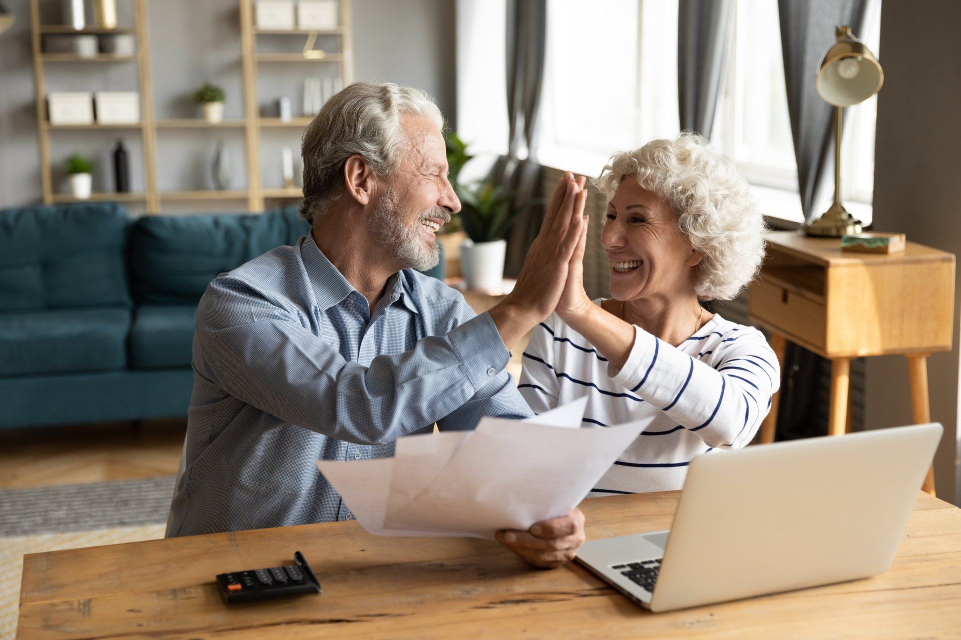 Smiling older couple giving a high-five, celebrating while reviewing paperwork at a table with a laptop.