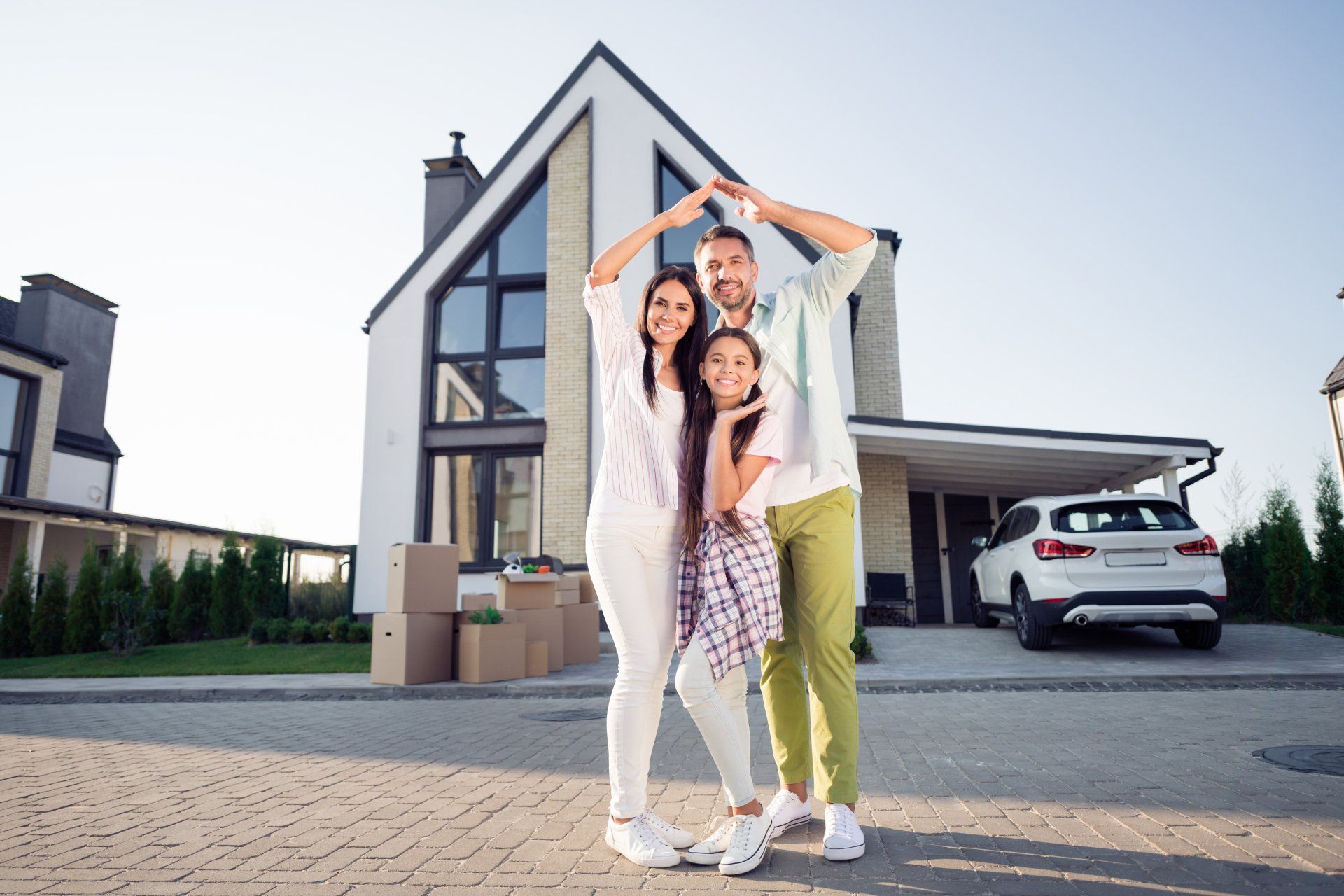 Family, arms raised in a roof shape, stands in front of their new home. Boxes and car visible.